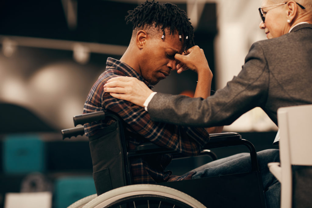 A worried young man in a wheelchair is comforted by a woman with her hand on his shoulder.